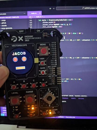 A photo of the supercon 2023 badge with vscode on a laptop screen behind it. On the badge's screen is a simple robotic face and my name, Jacob, in rainbow letters. Some Rust code is almost visible in vscode.
