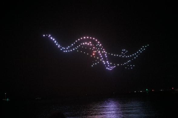A flock of drones form an image of the rainbow serpent Wargal fighting the ancestral crocodile Yondock over a smooth sea.
