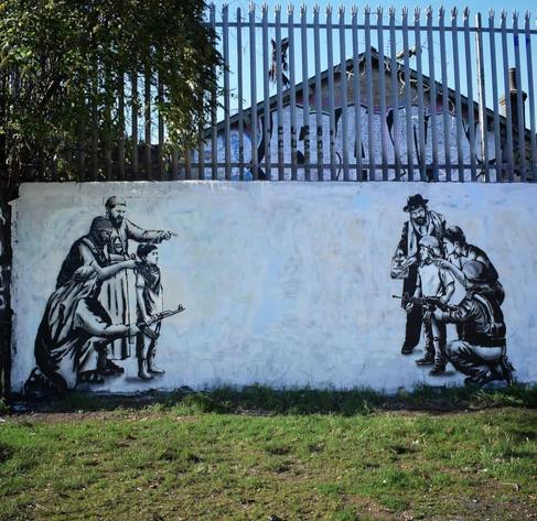 Streetartwall. On a white street wall with a small fence above, a critical mural about the future of the conflict between Israel and Palestine was sprayed. Designed only with black lines, it looks like a drawing. On the left, you can see a young boy with a Palestinian headscarf around his neck and a machine gun in his hand, being harassed by three adult men dressed in oriental clothing and holding a Koran. They talk at him and point to the right. To the right is a young boy wearing a helmet and holding a machine gun. Three Jewish-clad men holding a siddur are talking at him. The two children look unhappy.