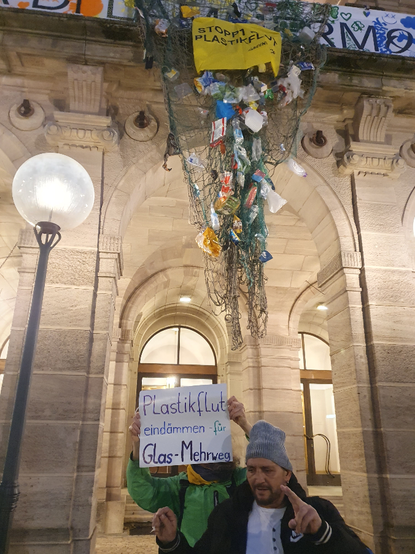 Netz mit Plastikmüll hängt vom Balkon des Opernhauses herab, darunter 2 demonstrierende Personen mit Plakat: Plastikflut eindämmen - für Glas-Mehrweg