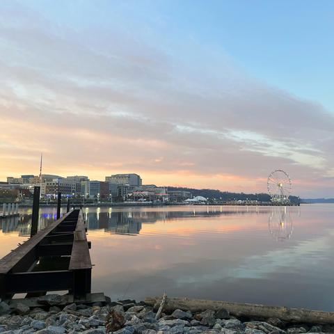 A view of the Gaylord National Resort and Conference Center and surrounding neighborhood in National Harbor, Maryland, from the north over the river. Thin clouds in the sky show hues of pink and yellow as they're illuminated by the rising sun out of frame to the left. The water is mostly still and reflecting the buildings and sky. A brown metal jetty extends in to the water on the left in the foreground.