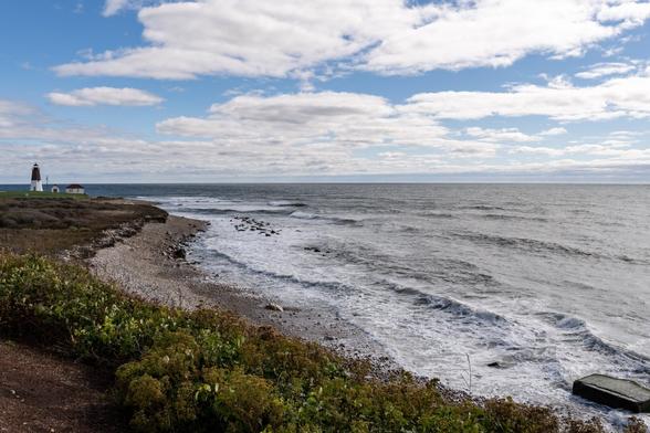 A color photograph taken during the day along a rocky coastline. In the foreground, a small line of coastal plants separates the vantage point from the rocks and water. A distance away, around the curve of the shoreline, a lighthouse stands, with two smaller buildings nearby. The lighthouse is white on the bottom half, and grey stone above. The light is oriented so it’s visible in the frame. 

The ocean is grey under a partly cloudy sky with swaths of blue, as an offshore gale had just passed.