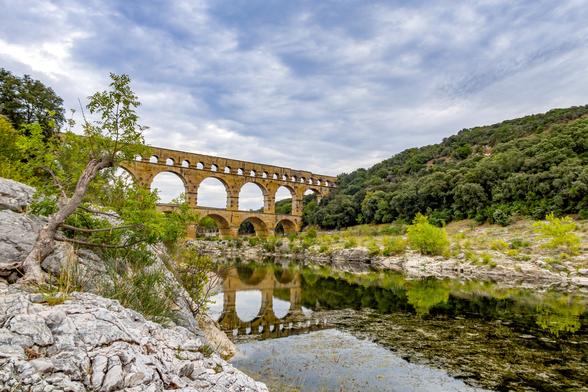 A view from a river bank of a tall and long Roman aqueduct spanning the river with a reflection of the structure on the still water. The aqueduct has three layers of arches supporting it two rows of large spans and top row of small spans. There are mottled clouds over the scene.
