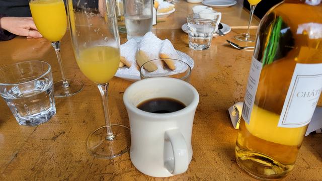 Photo of a cup of coffee, a champagne glass with a mimosa in it, and a plate of beignets covered with powdered sugar behind.
