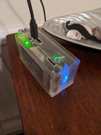 Pwnagotchi in 3d printed case, with usb cable sticking out of it, and a carabineer attached on the side loop. This angle shows the various LED signals emitting from the device through the translucent case. A peuter, oyster shaped tray in the background holds shells and a fake mustache