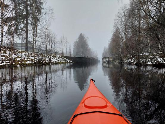 Kajakki lähestyy pitkää kanavaa. Rannat osittain lumiset. Sumuinen keli.