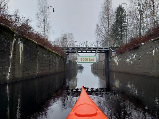 Kanavan ylittävä kävelysilta, josta roikkuu kyltti. Siinä lukee 3000 km eli kuluvan vuoden melontakilometrit tällä kohdalla. (Kyltti lisätty kuvaan jälkikäteen.)