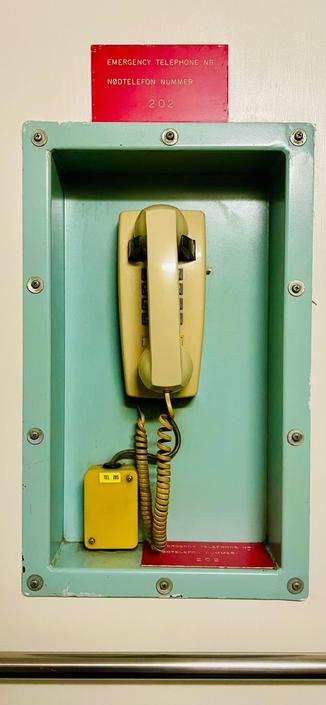 Old emergency phone in a mint green recess fitted to a vail white wall, with underneath a metal grab bar, and above a red sign indicating emergency telephone in English and Norwegian with the number 202.
