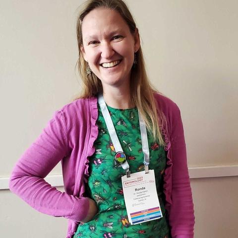 A woman with blond hair smiles as she wears a green shirt with a print of dozens of insects throughout, including flies, beetles, mosquitoes, wasps, true bugs, and more.
