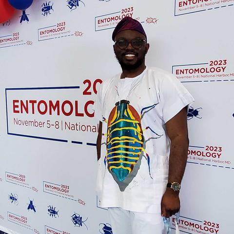 A man stands in front of an Entomology 2023 logo wall while wearing a white t-shirt with a single large beetle mostly iridescent green with many horizontal yellow stripes.