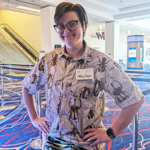 A person stands with their hands on their hips while wearing a beige collared button-down shirt featuring a repeated print of several beetles and other insects in black or dark brown.