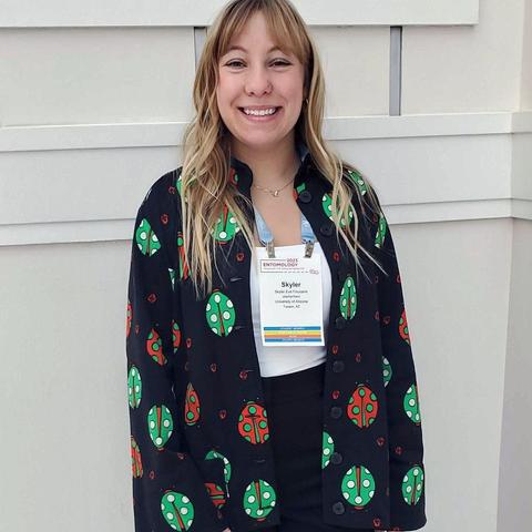 A woman smiles while wearing a large button-down shirt, unbuttoned over a white shirt, with a repeated print of ladybug-like insects, some red with green spots and others green with white spots.