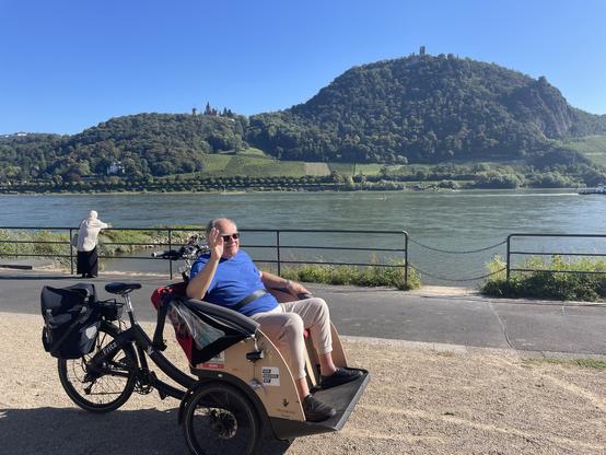 Ein älterer Herr sitzt vorne in einer Fahrrad-Rikscha und winkt. Im Hintergrund sind der Rhein, das Schloss Drachenburg und der bewaldete Drachenfels mit der Ruine zu sehen. Es ist ein klarer Tag im frühen Herbst mit blauem Himmel.