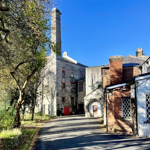 Bright blue sky with trees in the foreground and single storey red and grey coloured brick buildings to the side. In the background, a three storey grey brick mill building with a high rectangular brick chimney.
