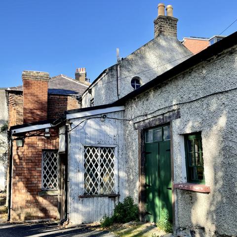 Pebble dash and red brick two storey and one storey buildings dappled in sunlight. Bright blue sky.