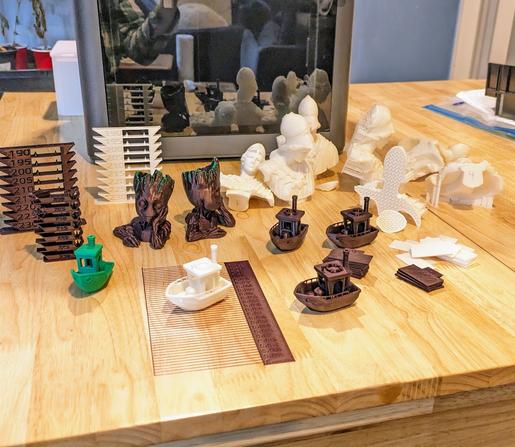 Multiple plastic figurines including boats, busts, and calibration objects are scattered about on a wood table in front of a 3D printer.