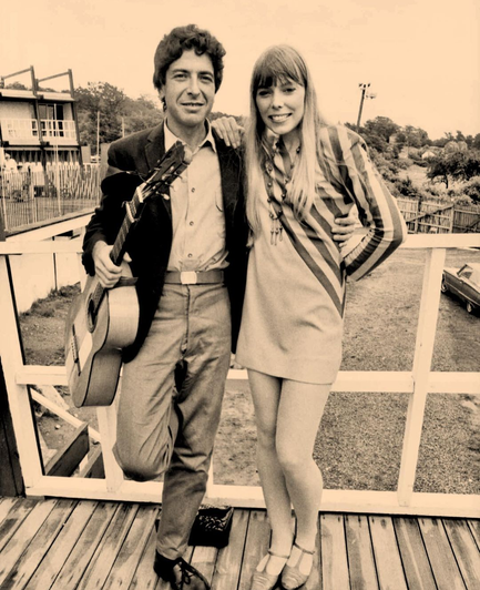 Sepia toned photo of a man with dark curly hair, dark blazer and an acoustic guitar stands arm in arm with a tall slim woman with long blonde hair, a short striped dress and a big smile
