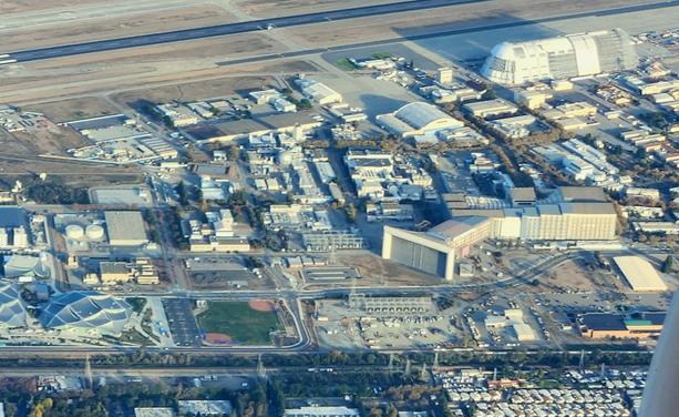 An aerial view of NASA Ames, featuring the wind tunnel complex, a large boxy building in the right half of the image with a horn shaped diagonal offshoot at one end