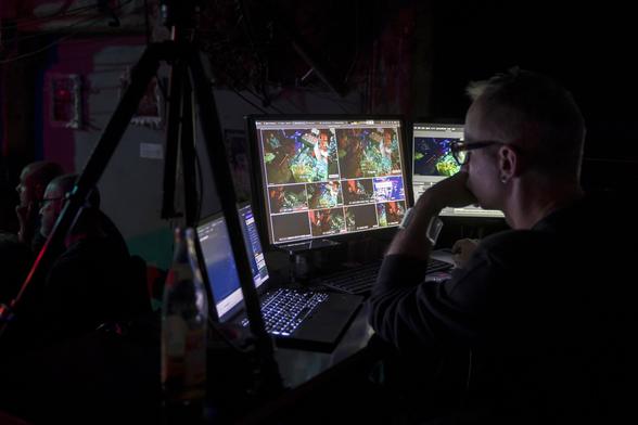 a white person behind two screens and a laptop in a dark seemingly editing live video stream.