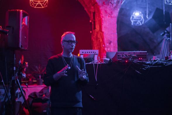 white person with glasses in front of a small stage seeminly explaining something. the place is lit with vivid color spectrum of red and pink