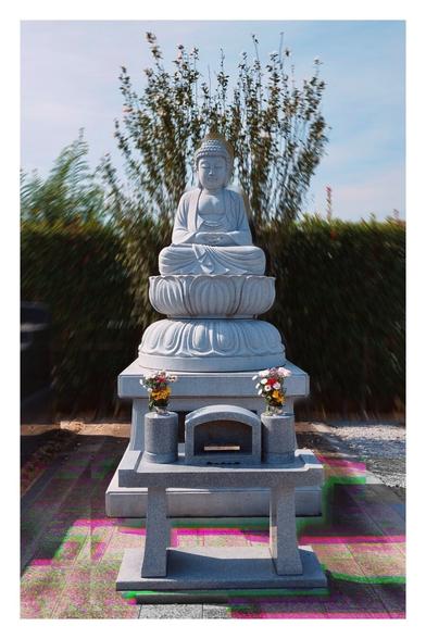 A picture of a Buddha sculpture, which is part of a shrine in a cemetery. It's all open air without a roof and made of marble. There is a shoulder height hedge behind it. I've edited the picture to give the greenery and the sky a blurred effect and the tiled ground an effect that adds red and green offsets of edges - it's hard to describe. The idea was just to weird it up, the picture by itself isn't that interesting.