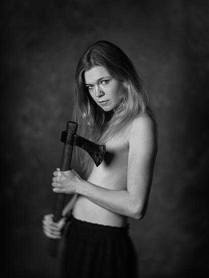 Junge Frau, die nur einen Rock trägt, hält eine Wikingeraxt in der Hand und schaut herausfordernd in die Kamera. Die Axt verdeckt die Brust.