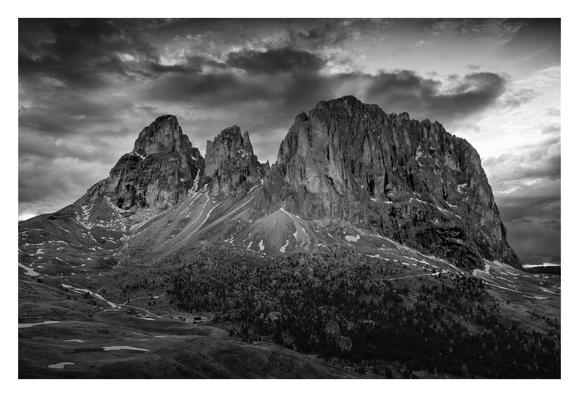 B&W photograph of Langkofel (3181m) just before sunset as seen from near Sella Pass in early June 2023. Moody, cloudy sky. Small patches of snow on the lower slopes. A piece of forest is gradually thinning out towards the talus fields below the rocky walls.