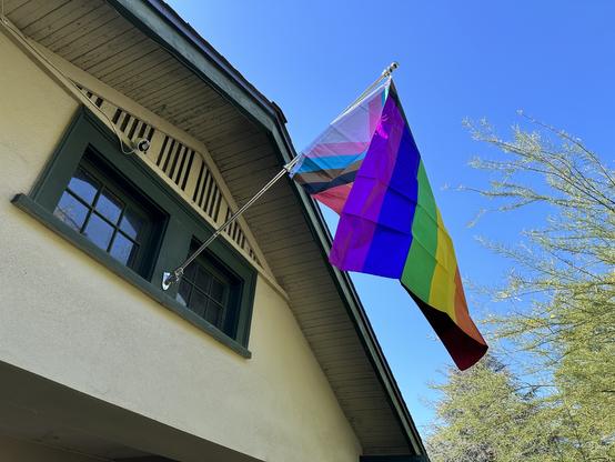 Folded over lgbtqia+ flag on a mast attached to the window frame in the gable above of our porch.