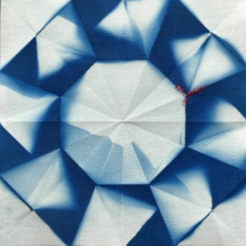 A dual octagonal geometric pattern in blue and white, on paper that shows the folds from previously being an origami form. There is red stitching mending small rips in the paper
