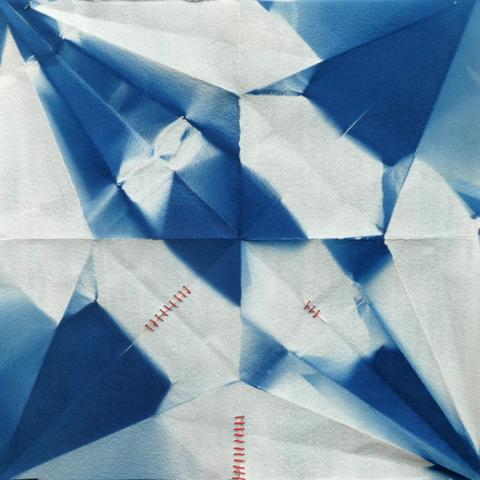 A mix of squares and triangles in an unbalanced geometric pattern in blue and white, on paper that shows the folds from previously being an origami form. There is red stitching mending small rips in the paper. This paper used to be an origami crane
