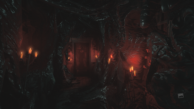 Zrzut ekranu z gry "Layers of Fear" przedstawiający bliżej nieokreślone pomieszczenie niemalże całkowicie zarośnięte czarną, bezkształtną masą, która pochłania i scala ze sobą wszystko, pulsując niczym żyjący organizm będący pasożytniczą naroślą. Z bezkształtnej masy wystają wysokie, mosiężne kandelabry w których palą się świeczki oświetlając pomieszczenie niepokojącym, czerwono-pomarańczowym światłem. Na wprost nas znajdują się obdrapane drzwi na których ktoś namalował czarną farbą staromodną wagę aptekarską.