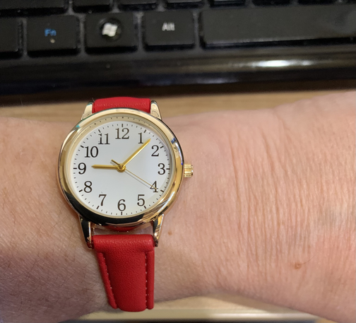 Analog watch on a wrist. Watch has a red strap. Watch face is white with gold trim. Large face and numbers. Time is 9:07
