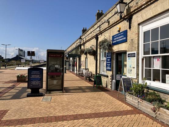 Similar to the previous picture, still on the station concourse but now looking along the length of the building. The outside of the Station Shop and Tourist Information (with flower tubs and signboards) is in the right foreground. The three long, straight terminal platforms are in the background, behind the phone boxes.  In the distance, beyond the platforms, some industrial buildings can be seen.
