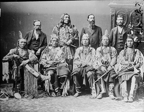 A group of men pose for a picture with some in Native American dress and long hair and others with shorter hair and western clothes