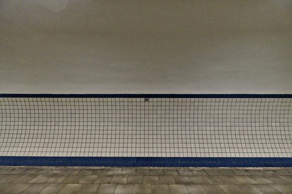 A minimalist image of a subway wall with small, square white tiles and a single blue stripe running along the bottom. A solitary black sign with white text is affixed to the wall above the blue stripe. The floor in front of the wall is paved with large square tiles, and the ceiling above is a smooth, plain surface.