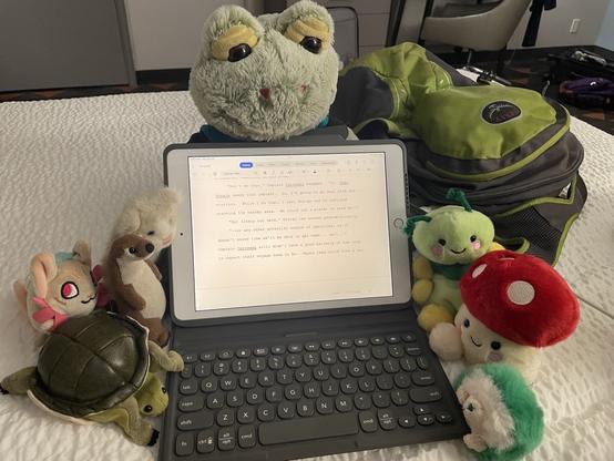 Photo of an iPad with a keyboard on a hotel bed with a ridiculous number of plushies clustered around it and a green backpack behind it. The plushies include a small otter, cat, turtle, sphinx cat, caterpillar, mushroom, green hedgehog, and large frog.