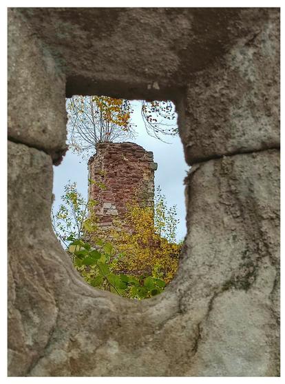Blick durch ein u-förmiges kleines Fenster aus dicken Feldsteinen. Durch das Fenster sieht man einen alten Turm aus Backsteinen, der teilweise mit herbstlich gefärbten Bäumen überwachsen ist.