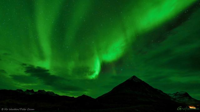 Lovely waves and ripples of green light in a dark sky, stars are visible too. The skyline is a jagged dark block containing a lovely pyramidal mountain, which is overtopped by the moving beams. A small building is lit and just visible on the right of the shot, lending it scale. Above a few small scattered clouds, many stars are visible, some even shining through the strong beams of aurora.