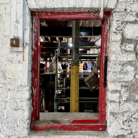 A red framed glassless window in an internal whitewashed brick wall shows machinery and scaffolding in another room.
