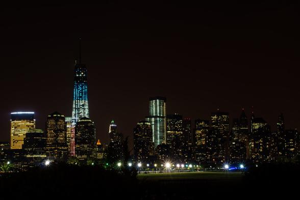 A color photograph taken at night of the lower Manhattan skyline. The lit shape of One World Trade looms over the other buildings. In the foreground, the manicured lawn of a New Jersey park, brightly lit, is visible. The Hudson River can’t be seen, somewhat erasing the distance to the buildings.