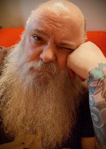 Me, a big-bearded, chunky, bald, biker-Santa looking cis-dude sitting on my boyfriend's sofa, squinting at the camera having woken myself up, coughing, again.