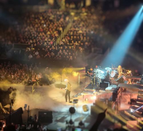 Slightly fuzzy, side-on image of an aging guitarist jamming in front of a man on a drum set. The spotlight is on the drummer. Past them, every seat in the stands is full.