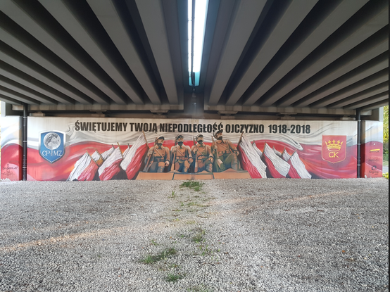 Mural pod wiaduktem. Szeroki obraz który w tle ma flagę bialo czerwoną.
U samej góry napis: Świętujemy twoją niepodłegłość Ojczyzno 1918-2018.
Poniżej napisu, na środku - cokół na którym stoi czterech żołnierzy.
Po prawej i lewej stronie od żołnierzy, fłagi Polski. Na zewnatrz po lewej, herb  z napisem CPMZ - Centrum Przygotowań do Misji Zagranicznych.
Na zewnątrz, po prawej, herb Kielc.