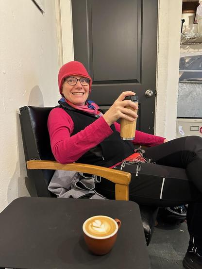Smiling white woman wearing glasses, a pink hat, pink long sleeve shirt with a black vest over it, brightly colored scarf, and black pants holding a tall bamboo coffee go mug in her hand while she sits in a black chair. On the black table in front of her, a cappuccino with a frothy leaf of latte art in the top foam.