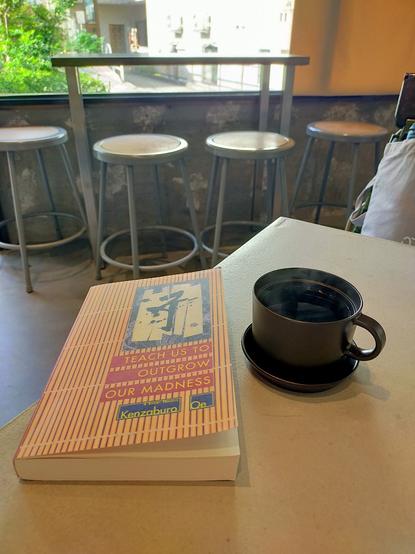 The photo is of a table inside a cafe. The paperback book is designed to look like a bamboo wall with kanji calligraphy in the upper right corner. To the left is a black mug & saucer of black coffee. In the distance is a counter against a window with 4 metal stools