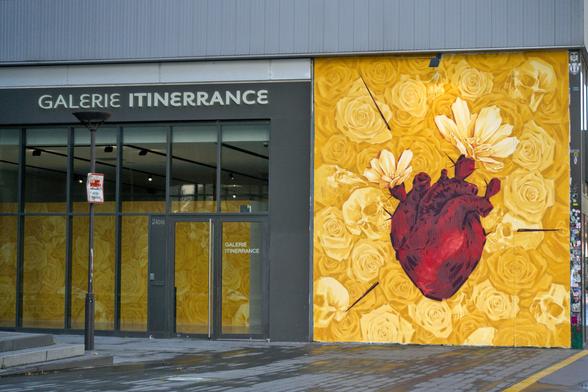 Façade de la galerie Itinerrance. Sur la droite, le mur est recouvert d'une œuvre d'Inti représentant un fond de roses et de cranes jaunes avec, au centre, un cœur d'où sortent des épines et des fleurs. À gauche de la peinture murale, une surface vitrée indique « Galerie Itinerrance ». À travers les vitres, on peut voir un mur entièrement peint de roses et de cranes jaunes.