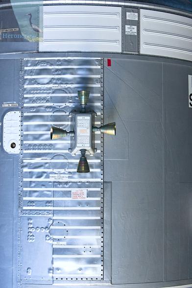 A photograph of part of a gray metal cylinder with white stripes in places. In the center, there is a four-sided box with four rocket engine nozzles sticking out of each of the four sides.
Small labels with text and graphics are on and around the box.
