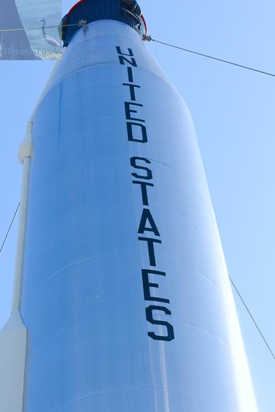 An outdoor, daylight photograph of a shiny, silvery metallic rocket body of a cylinder topped by a conical section. The words “United States” are written vertically on the rocket’s body. The sky is a light blue and is reflected by the silvery skin of the rocket.