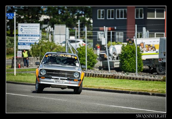 A Mk2 Escort Drifting around a tarmac corner of a street closed for racing, facing towards the viewer as the photographer was positioned on the inside of the corner. It has wide rally flares which are in orange, the rest of the car is in silver. It is being counter-steered way from the viewer to continue the drift around the corner and carry on. Industrial area fences and buildings are out of focus in the background.