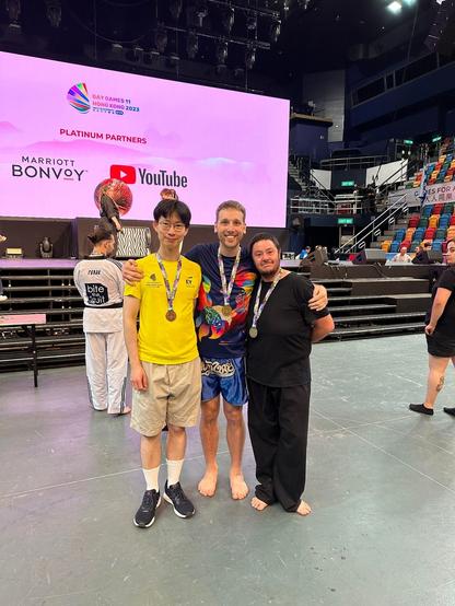 Picture of 3 people wearing medals in a stadium. On the left is a young east Asian man wearing a bronze. He's in street clothes. In the middle is a white masculine person wearing a gold. He's barefoot in muay thai shorts and a Gay Games shirt. On the right is a light skinned Aboriginal man wearing a silver. He's barefoot in a black gi. They are smiling and embracing.